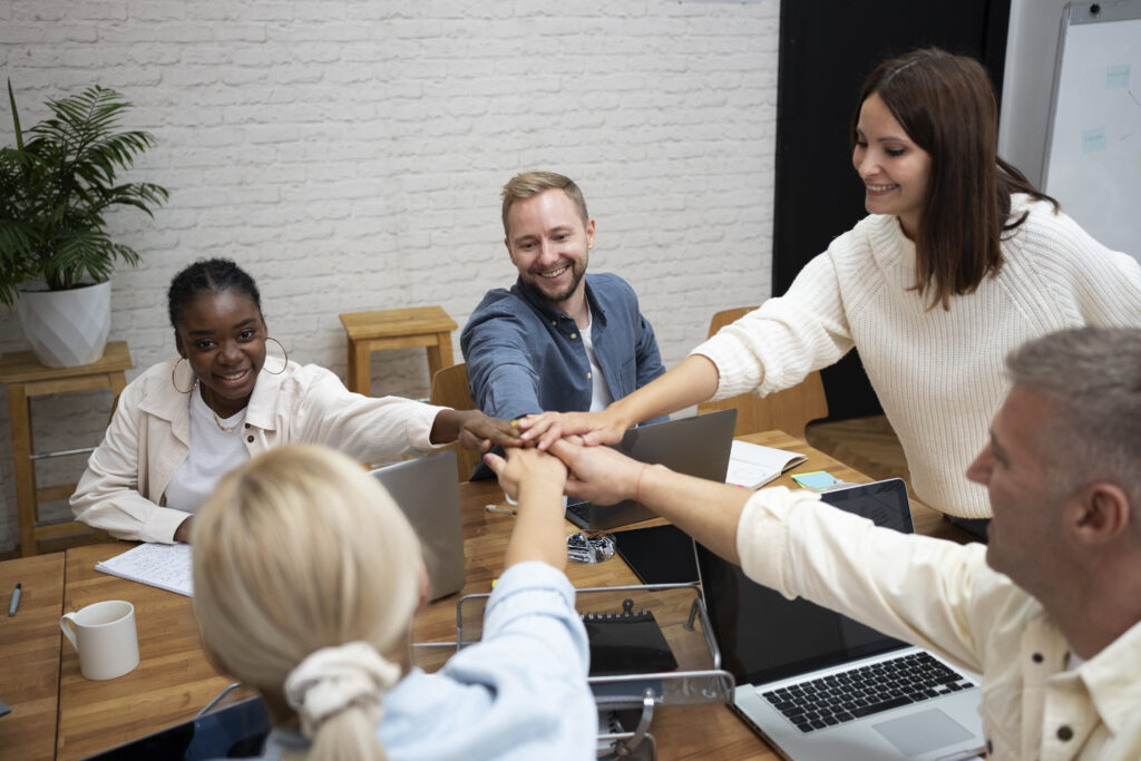 Equipe diversa reunida em um ambiente de escritório, unindo as mãos no centro da mesa em um gesto de colaboração. A imagem representa a superação da cultura de silos nas empresas, com profissionais engajados trabalhando juntos de forma integrada. participantes do workshop de primeira liderança