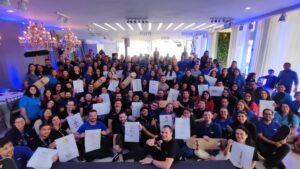 Imagem de um evento de Team Building de fim de ano, com um grande grupo de funcionários sorrindo enquanto seguram pranchas de skate personalizadas e desenhos. O ambiente é descontraído e festivo, com decoração iluminada e um clima de celebração de propósito. A equipe está unida, refletindo o fortalecimento e engajamento da empresa para o novo ciclo.